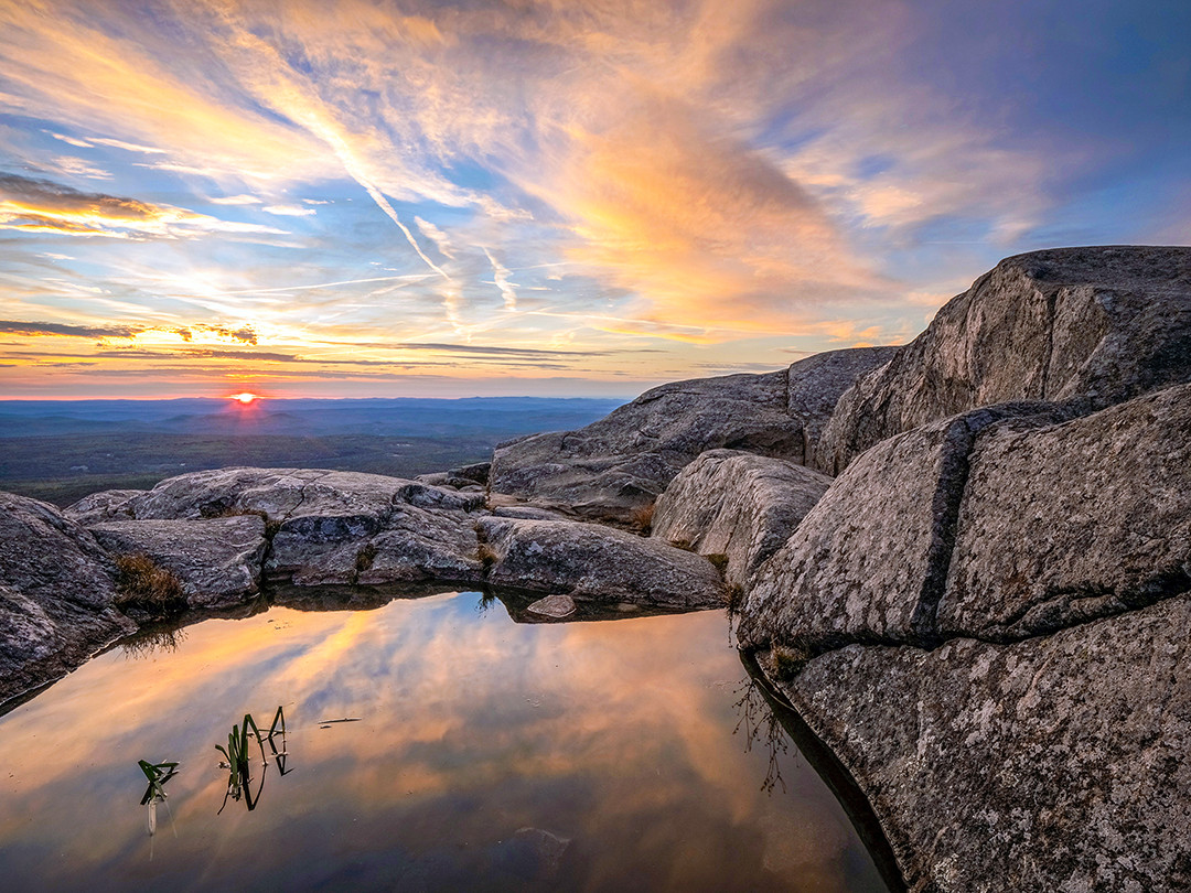 The setting sun reflected in a rain-fed pool, at the summit of Mount Monadnock in New Hampshire.⁠
⁠
At an elevation of 3,165 feet (965 m), Mount Monadnock is the highest mountain in southern New Hampshire, towering over 1,000 feet (305 m) higher than any other peak within 30 miles (48 km). Although it is shorter than many of the peaks in the not-too-far-away White Mountains, Mount Monadnock is one of the most frequently climbed mountains in the world, given its proximity to major cities such as Boston, and its high reward in terms of views given a fairly moderate hike.⁠
⁠
The White Dot and White Cross Trails are the most common points of access to the summit of Mount Monadnock, coming in at just under 4 miles (6.4 km) round trip, climbing 1,774 feet (541 m) of elevation. While this is no minor task, relatively speaking this is a rather short hike, given the incredible views up top.⁠
⁠
Due to the bald, granite-covered summit, there is essentially no soil or drainage layer, so any rain, snowmelt, or condensation tends to linger around a bit longer than on usual surfaces. Given the recent rain in the days leading up to this hike, it made sense that there were many pools on the summit, providing me with a variety of surfaces to use for reflections, mirroring the amazing sunset taking place in the sky on this recent autumn evening.