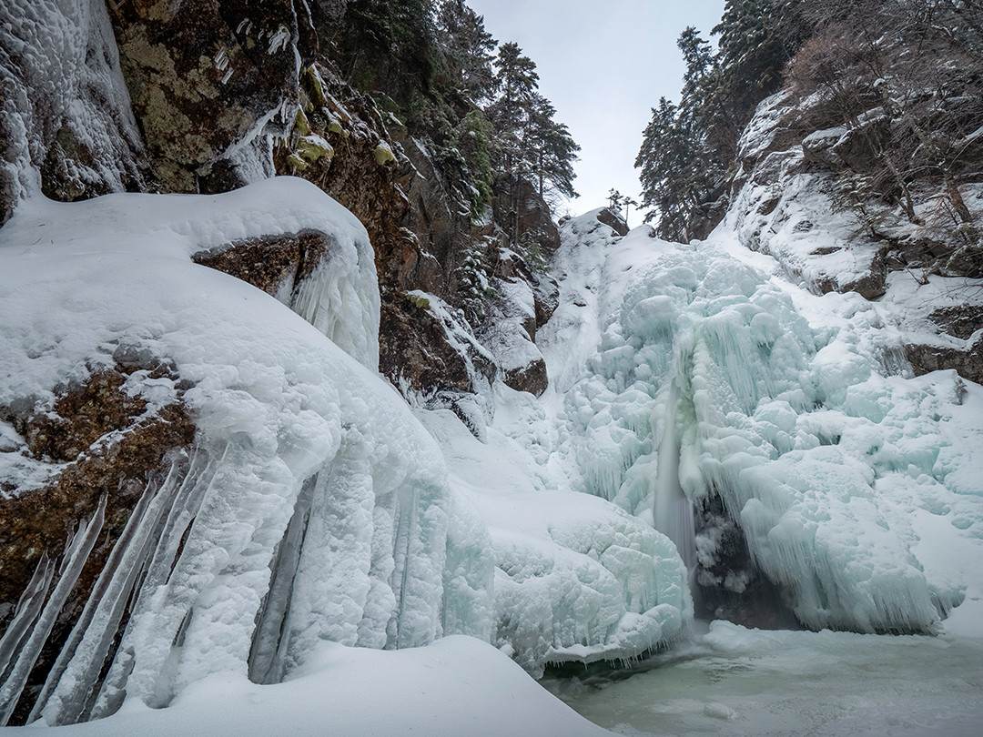 An almost entirely frozen Glen Ellis Falls, one snowy morning in White Mountain National Forest, New Hampshire.⁠
⁠
Glen Ellis Falls is a 64-foot (19.5 m) tall waterfall located in Pinkham Notch, a mountain pass in the White Mountains of New Hampshire. Beautiful all year round, its appearance dramatically changes during periods of sustained winter cold, allowing it to almost entirely freeze over. This effect is further enhanced during periods of recent or ongoing snowfall, as was the case on this morning.⁠
⁠
Spray from the plunge zone repeatedly coats the basalt and granite walls, resulting in bulbous ice layers building outward, rather than the single solid sheets of ice other waterfalls might display. Because this waterfall sits in a heavily shaded notch of the mountain, with limited sun exposure and frequent cold air pooling, it is more likely to freeze over completely than many other waterfalls found in the White Mountains.⁠
⁠
This scene was beautiful to witness in person, as it snowed softly throughout the entire time spent photographing here. The fresh snow covered the layers of icicles present, and between the sound insulation provided by the snowpack and the far less furious roar of the frozen waterfall, the scene felt far more peaceful and serene than it would during the warmer months.