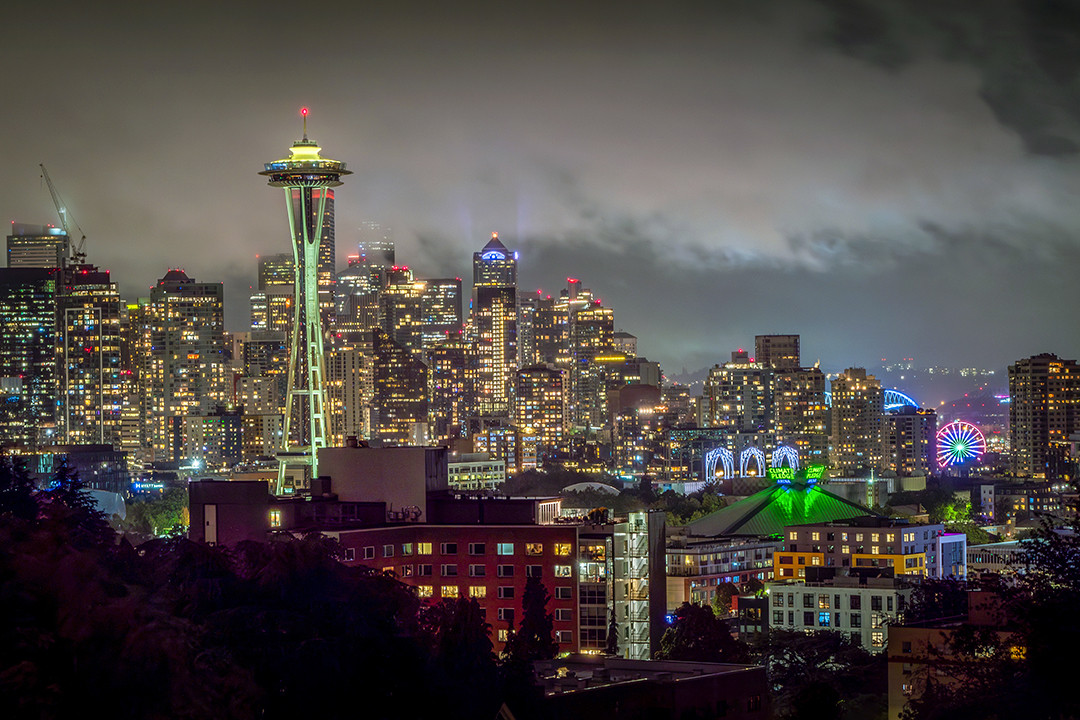 The glowing Seattle skyline rises beneath a fog-laced night sky, viewed from Kerry Park.⁠
⁠
On my trip to the Pacific Northwest, I spent a total of one brief night in Seattle, and I knew I wanted to photograph the skyline. Kerry Park is considered by most as the classic iconic place to do so, as it is located on the south slope of Queen Anne Hill, giving it a direct and elevated line of sight at downtown Seattle. Prominently featured from this vantage point is, of course, the Space Needle, with the Seattle Great Wheel peeking through a gap in some of the buildings to the right-hand side.⁠
⁠
The fog-laced sky is typical of the atmosphere here, when a marine layer of cloud cover forms as cool Pacific air moves inland, condensing at low elevations. Not only do these clouds mingle with the skyscrapers themselves and create great depth and visual contrast, but their low level allows the lights of the city to reflect off of them, amplifying their brightness. It was slightly raining on this evening, which, when combined with the atmospheric fog, resulted in my brief night here being classic Seattle, matching the classic nature of this viewpoint.