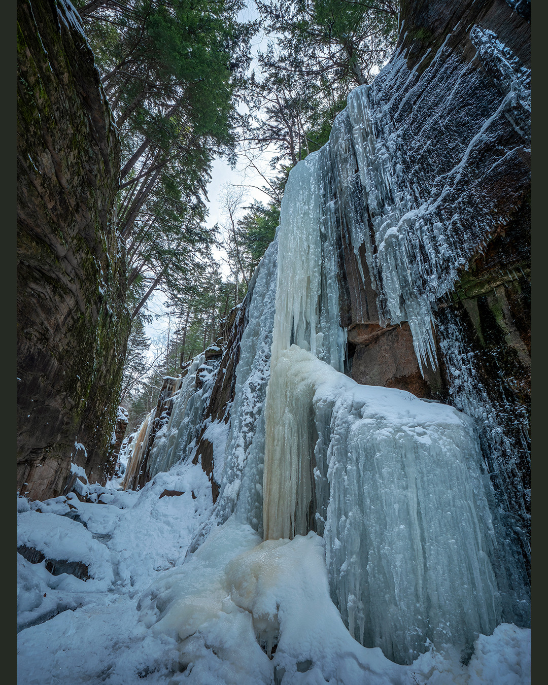 A frozen waterfall clings to the towering granite walls of Flume Gorge, on a subzero morning in the White Mountains, New Hampshire.⁠
⁠
I took this photograph over the winter on a few days trip through the White Mountains, in New Hampshire. This was on the first day, and temperatures were hovering around 0 F (-18 C) this morning, which coupled with the wind made for a blustery winter stroll to get here.⁠
⁠
This waterfall is quite popular with ice climbers, so to grab a photo or two before they arrived meant showing up shortly after sunrise. This provided a good 45 minutes of shooting before the started to show up. The reason this is a popular ice climbing spot is partly due to how thick and solid these ice curtains are. Unlike bigger waterfalls with higher discharge, these smaller cliffside ones freeze more completely due to their slower flow and volume. Also aiding the freezing is the Flume Gorge itself. The narrow gorge traps cold air, while restricting the sunlight which can reach below.