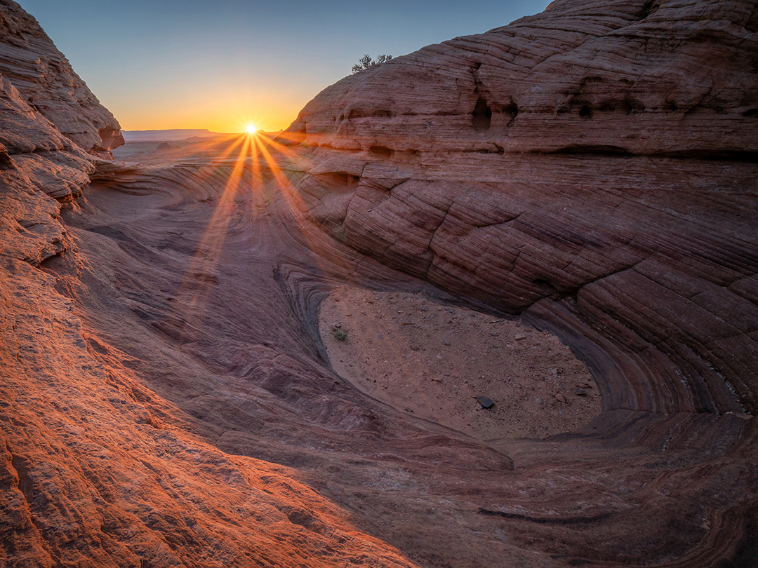 The rising sun crests over the edge of a wave-sculpted sandstone bowl early in the morning near Page, Arizona.⁠
⁠
On a recent trip to Page, Arizona, I wanted to make sure I took advantage of as many photographic locations as this region had to offer, which is quite a few. Most prominently known for Horseshoe Bend and Antelope Canyon, the sandstone wonders don’t stop there. As I was searching for other spots to photograph before embarking on this trip, something referred to as “The New Wave” caught my attention. I have always wanted to visit the actual Wave, about two hours farther west from Page, so surely the New Wave would be something worth visiting.⁠
⁠
Located along the Beehive Trail in Glen Canyon National Recreation Area, finding the actual New Wave proved to be fairly difficult. After about an hour of climbing around the sandstone mounds and bowls in this area, I finally arrived at the New Wave, which, coincidentally, is not where I took this photograph. This was the afternoon before I captured this image, and I was looking for a specific area where I could photograph the rising sun. The New Wave did not provide an appealing way of achieving this, so I continued scouting, eventually finding this particular wavy bowl about 500 feet away, sporting a pretty similar aesthetic.⁠
⁠
I made note of the spot and ventured back the following morning, about an hour before sunrise. I picked my composition and waited for the sun to pass over the top of the sandstone walls, illuminating the rock face with its warm glow and signaling the start of a new day.