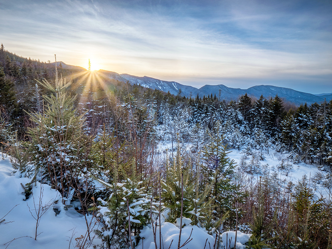 The setting sun peeks over the Sandwich Range, at the end of a wintry day in the White Mountain National Forest, New Hampshire.⁠
⁠
Unlike the sharper, more alpine peaks of the Presidential Range of the White Mountains, the Sandwich Range has more rounded summits and longer ridgelines, due to this being an older mountain range, shaped over millions of years by heavy erosion and repeated glaciation. The most prominent peaks here are Mount and East Osceola, and the forested bowl in the midground of this photograph drains toward the Pemigewasset River. The foreground is filled with young balsam firs and red spruce saplings, short in size and barely able to push up through the thick snow here.⁠
⁠
This photograph was taken from the Pemigewasset Overlook, one of the four major overlooks featured on the Kancamagus Scenic Byway. I stopped at all four on this day, and while the other three were fully plowed of snow, this one, for whatever reason, was not. Which, being in an all-wheel drive vehicle, proved to not be a major issue. The same could not be said for a couple of the other visitors who happened to pull into this parking lot on this afternoon. Of the upwards of an hour spent at this overlook, the majority of it was spent listening to the whirring wheels and laborious manpower efforts of the passengers trying to push their vehicles back out of the thick snow this parking lot was composed of.