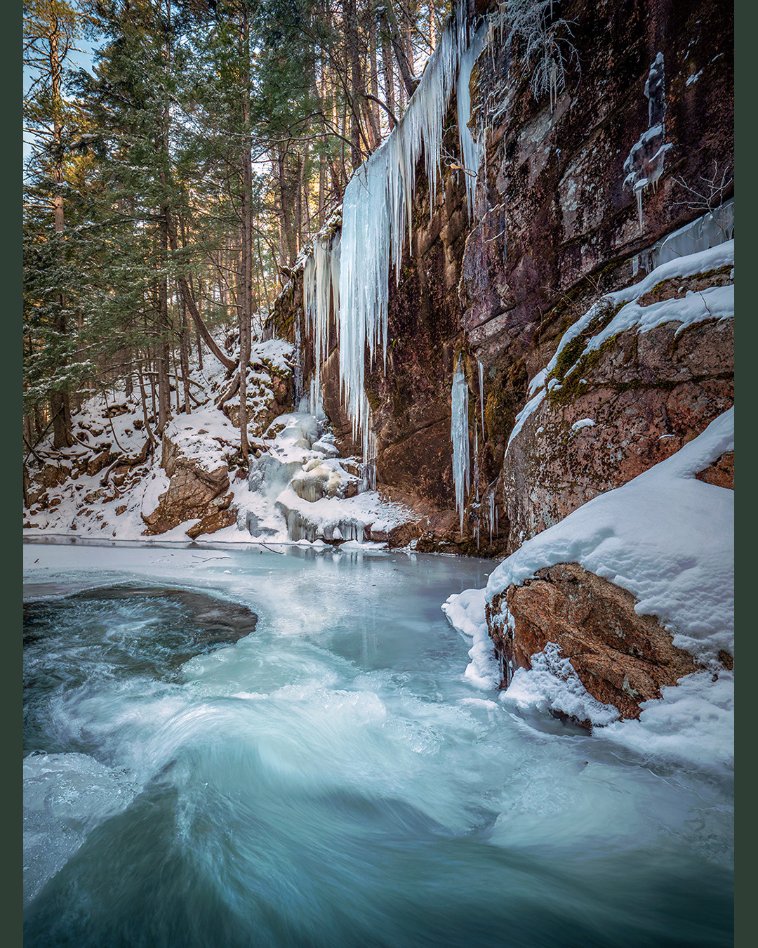 The frigid waters of Sabbaday Brook flow alongside their frozen counterparts on a winter afternoon in the White Mountains of New Hampshire.⁠
⁠
The few days spent in the White Mountains this past winter were quite frigid, with the temperature being around 5°F (-15°C) on this afternoon. Even in the depths of winter, where the water temperatures are extremely cold, the fast-moving, oxygen-rich water resists freezing completely over. The parts which are calmer and slower-moving develop surface ice and anchor ice formations. The swirling water in the foreground and the area where it plunges into remain unfrozen, while the water around the perimeter freezes over.⁠
⁠
This photograph was taken just below Sabbaday Falls, literally behind my back as I took this photo facing away from it. The curtain icicles forming along the rock face occur where seepage or splash freezes gradually over time. I placed one of the fully extended legs of my tripod directly in the rushing water, with the other two (much shorter) legs on the ice shelf to the side, with the camera only a few inches above the rushing water. This allowed me to use the flow of the water as a leading line into the plunge pool and icicle display.